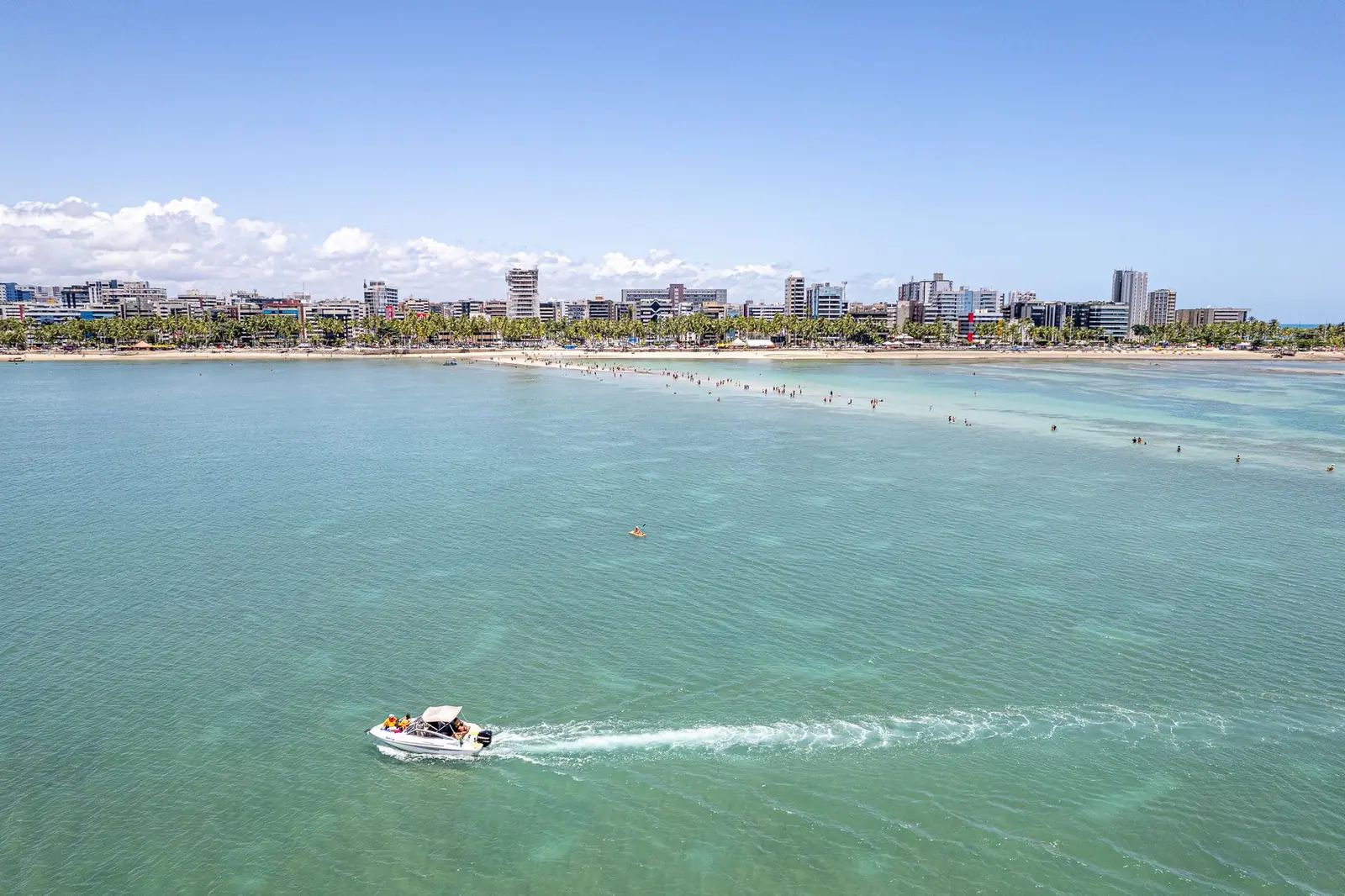 Lancha cruzando o mar verde de Maceió com a orla de prédios ao fundo